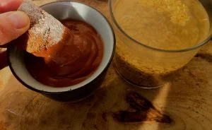 chocolate a la taza junto a un vaso con grano de avena a remojo y una mano untando bizcocho en el chocolate